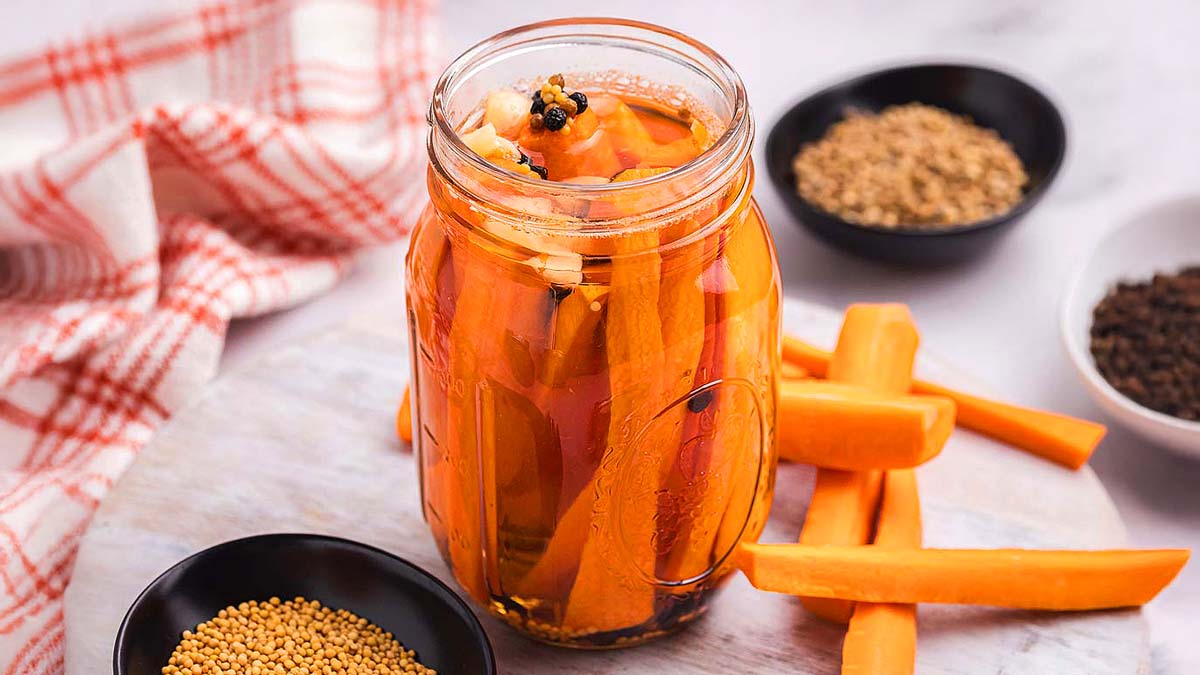 A glass jar filled with pickled carrot sticks sits on a round board, surrounded by loose carrot sticks and small bowls of spices.