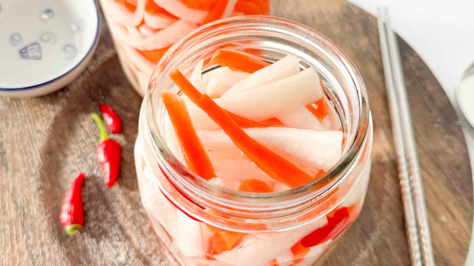 A glass jar filled with pickled carrot and daikon strips sits on a wooden surface next to red chili peppers, chopsticks, and a small dish.