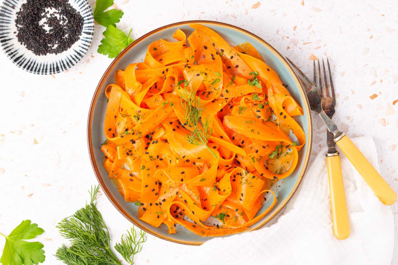 A plate of carrot ribbon salad garnished with black sesame seeds and fresh herbs, with a fork, knife, and herbs on the side.