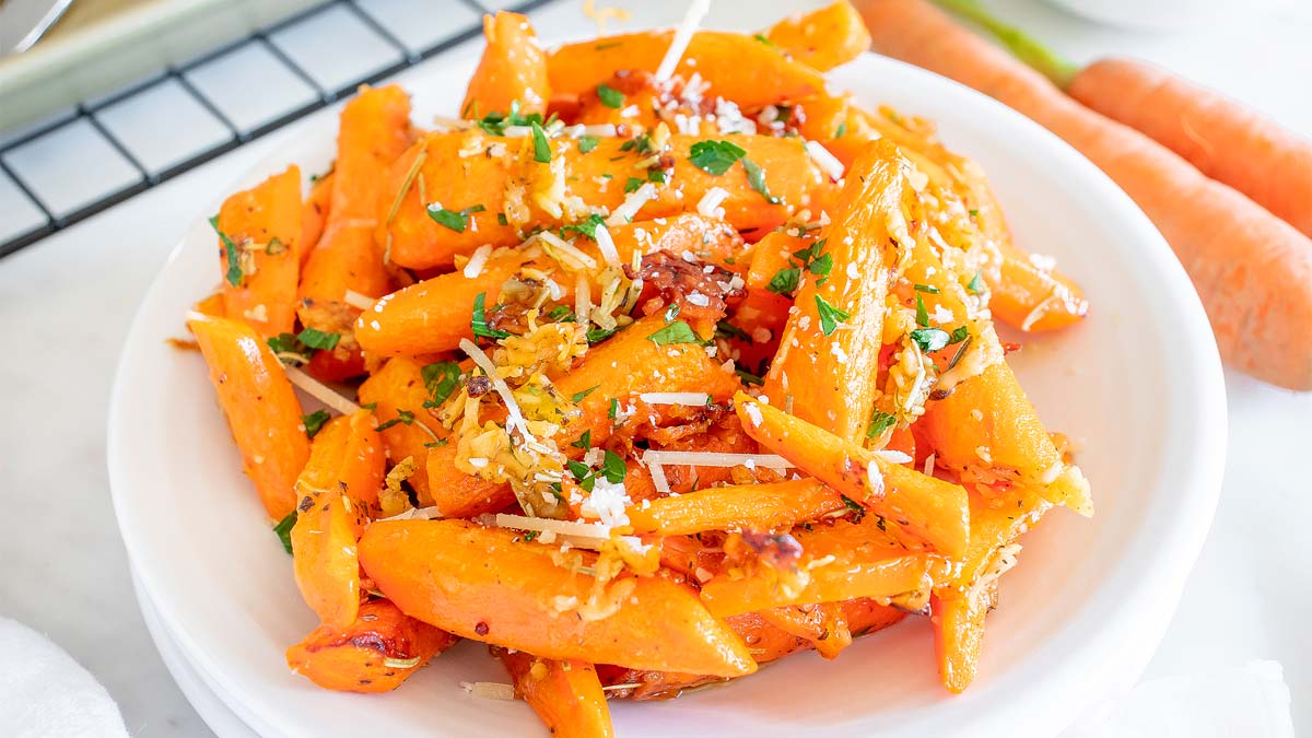 A white bowl filled with roasted carrot sticks garnished with chopped herbs and grated cheese, placed on a white surface.
