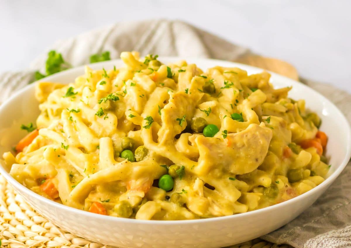 A bowl of creamy pasta with pieces of chicken, green peas, and carrots, garnished with chopped parsley.