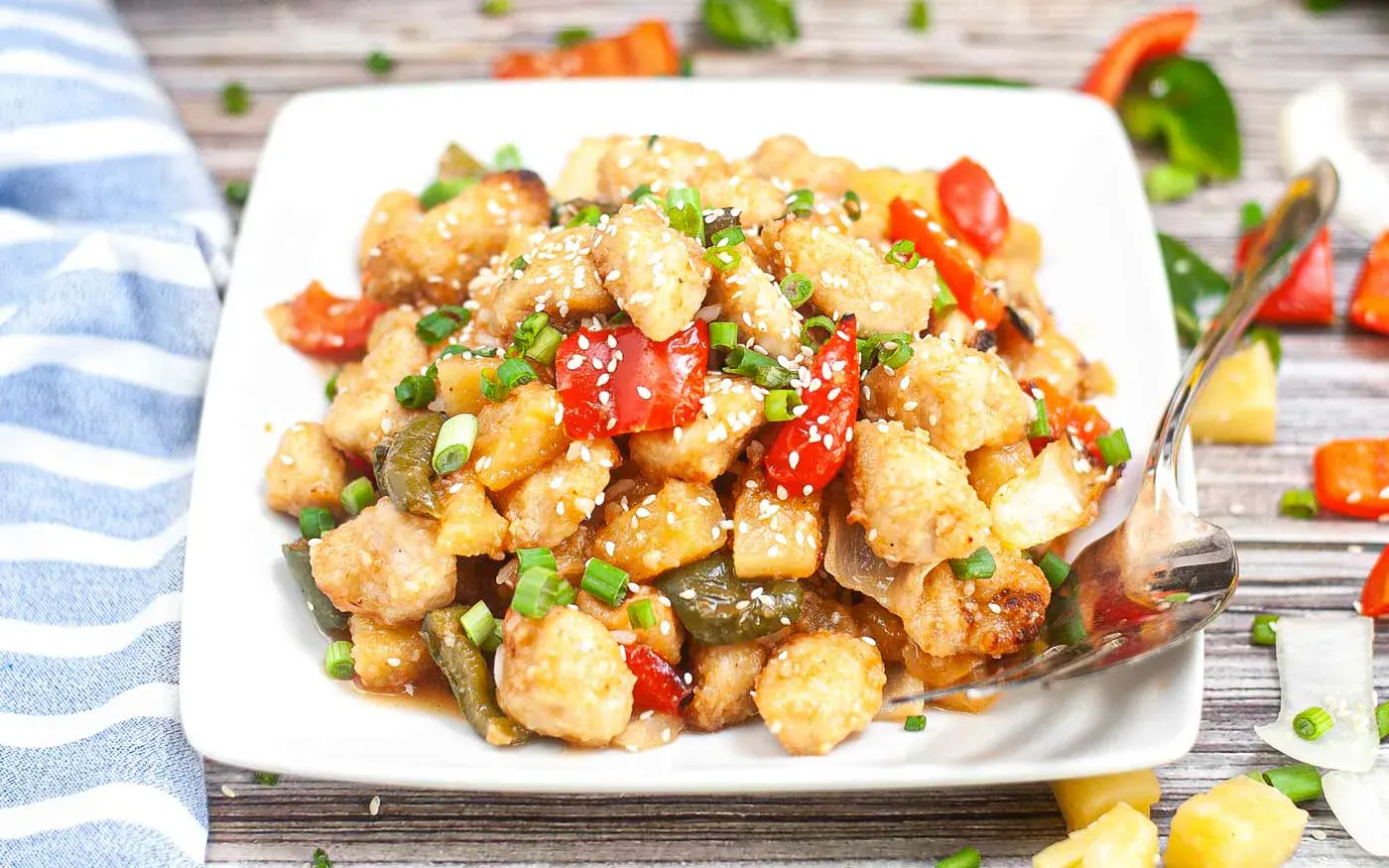 A white plate of sweet and sour chicken with diced vegetables, pineapple chunks, and sesame seeds, garnished with chopped green onions. A fork rests on the plate’s side.