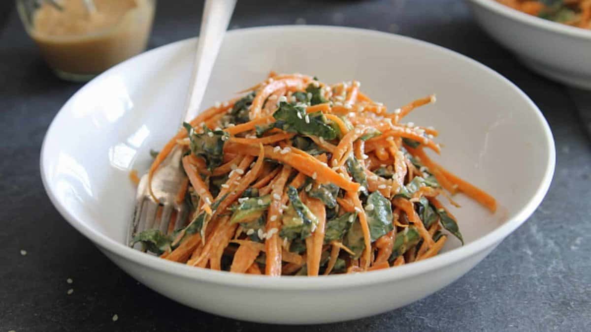A white bowl filled with shredded carrots, greens, and sesame seeds, with a fork resting inside.
