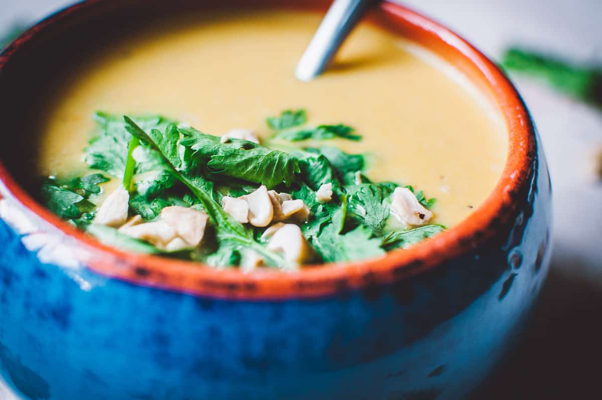 A bowl of creamy soup garnished with fresh cilantro and chopped nuts, with a spoon resting inside the bowl.