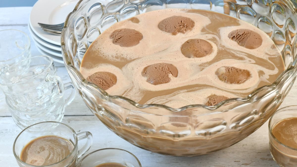 A glass punch bowl filled with creamy brown punch and scoops of chocolate ice cream, surrounded by glass cups and stacked plates on a white wooden surface.