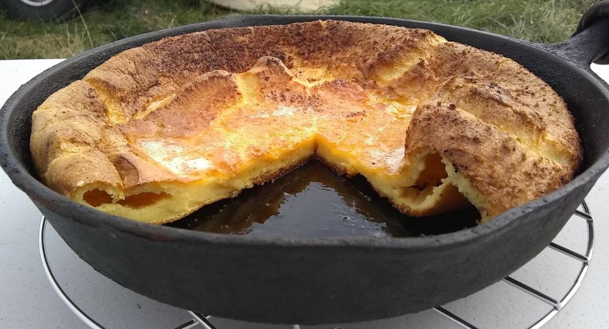 A baked Dutch baby pancake in a cast iron skillet with a wedge missing, showing its golden, puffy texture and browned edges, resting on a wire rack.