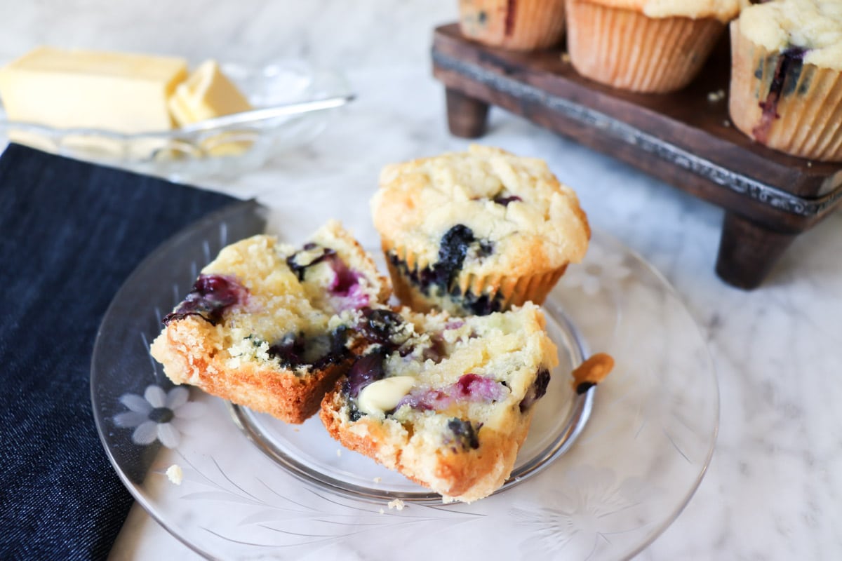 A blueberry muffin split in half on a glass plate with a pat of butter, with more muffins and a butter dish in the background.