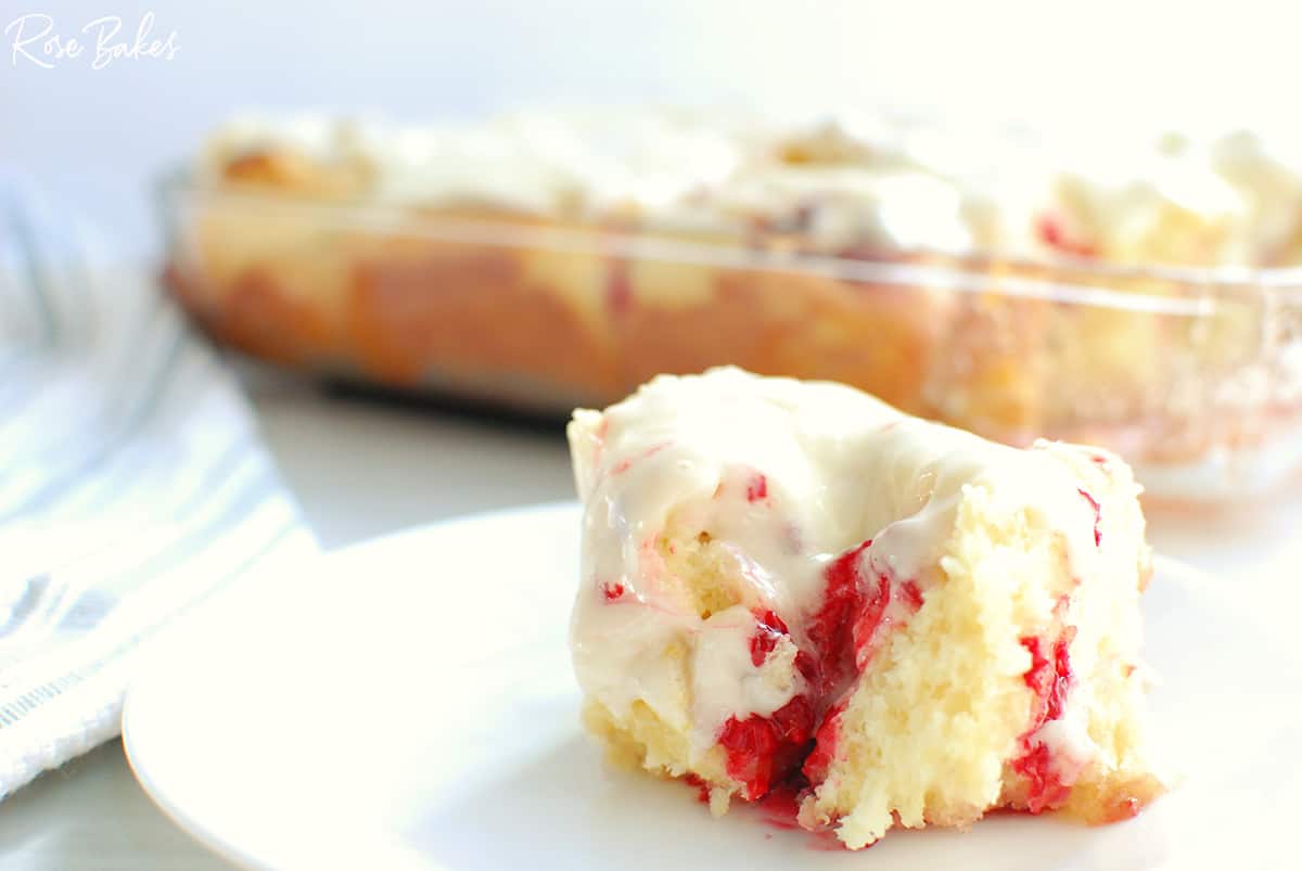 A piece of frosted cherry coffee cake sits on a white plate, with a glass baking dish containing the rest of the cake in the background.