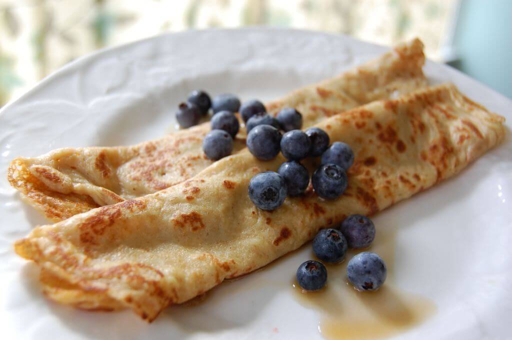 Two rolled crepes on a white plate topped with fresh blueberries and syrup.