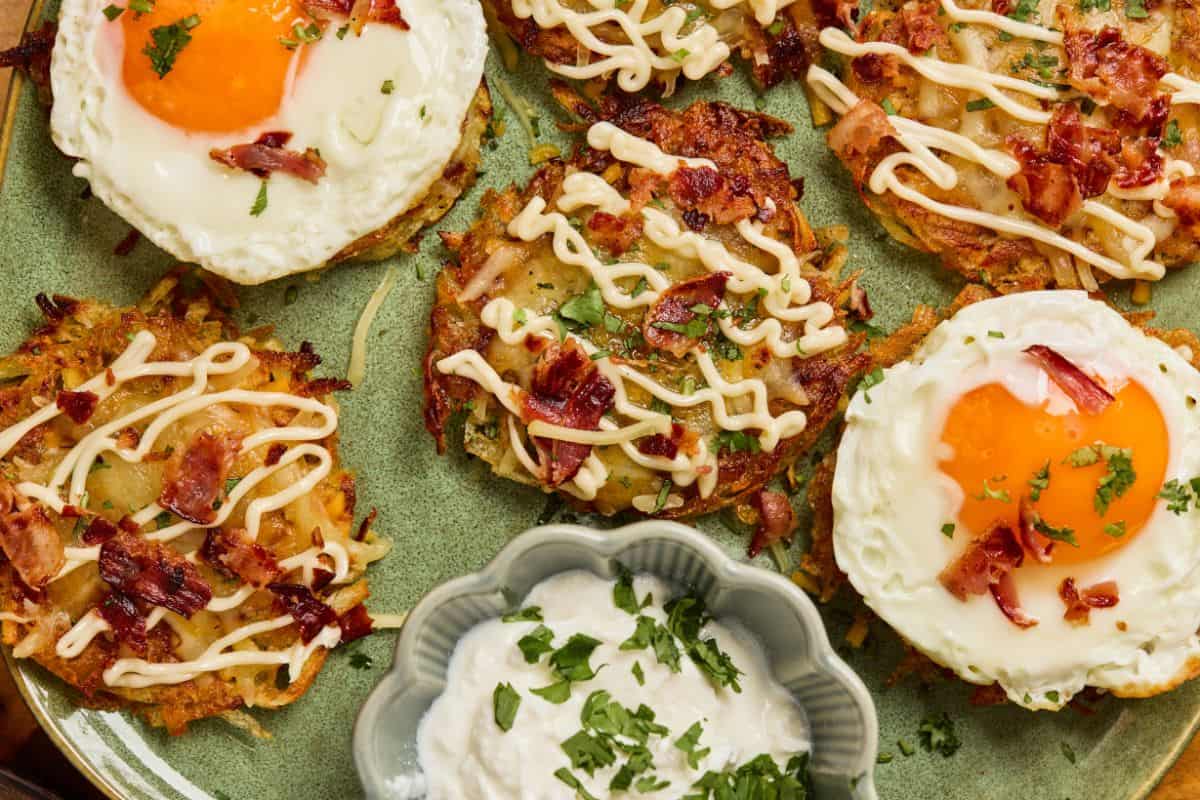 Potato pancakes topped with fried eggs, bacon bits, and drizzled sauce, served with a bowl of sour cream garnished with chopped herbs.