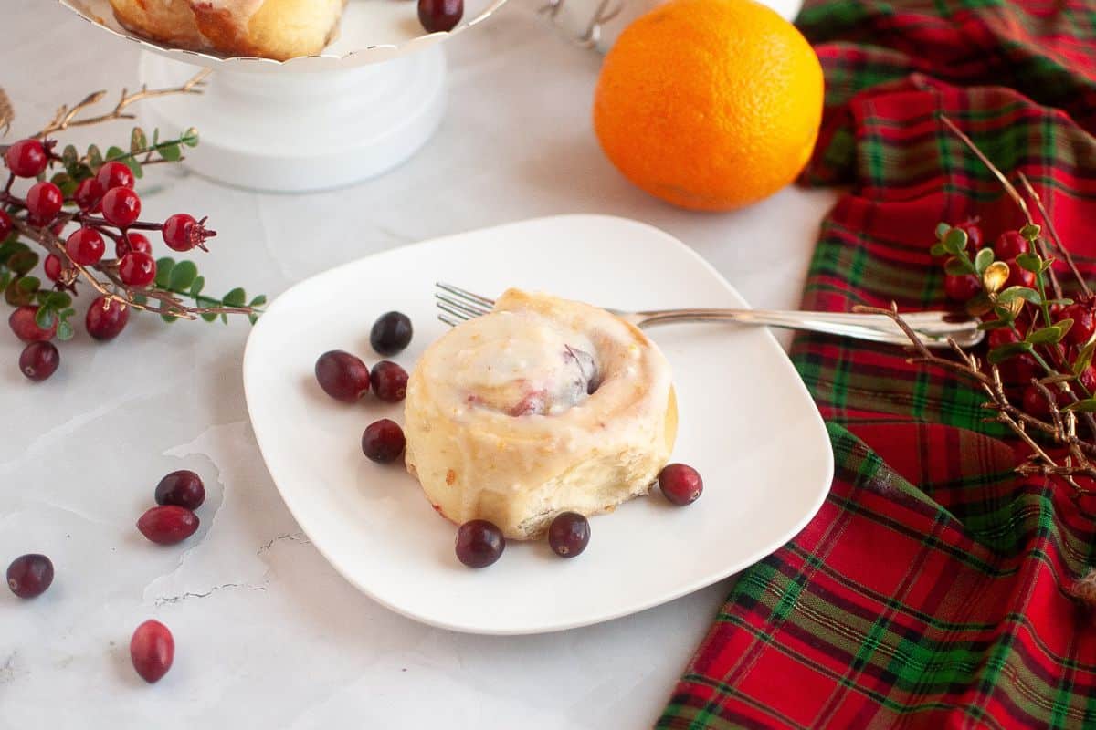 A glazed cinnamon roll with cranberries on a white plate, garnished with fresh cranberries, next to an orange and a red-green plaid cloth.