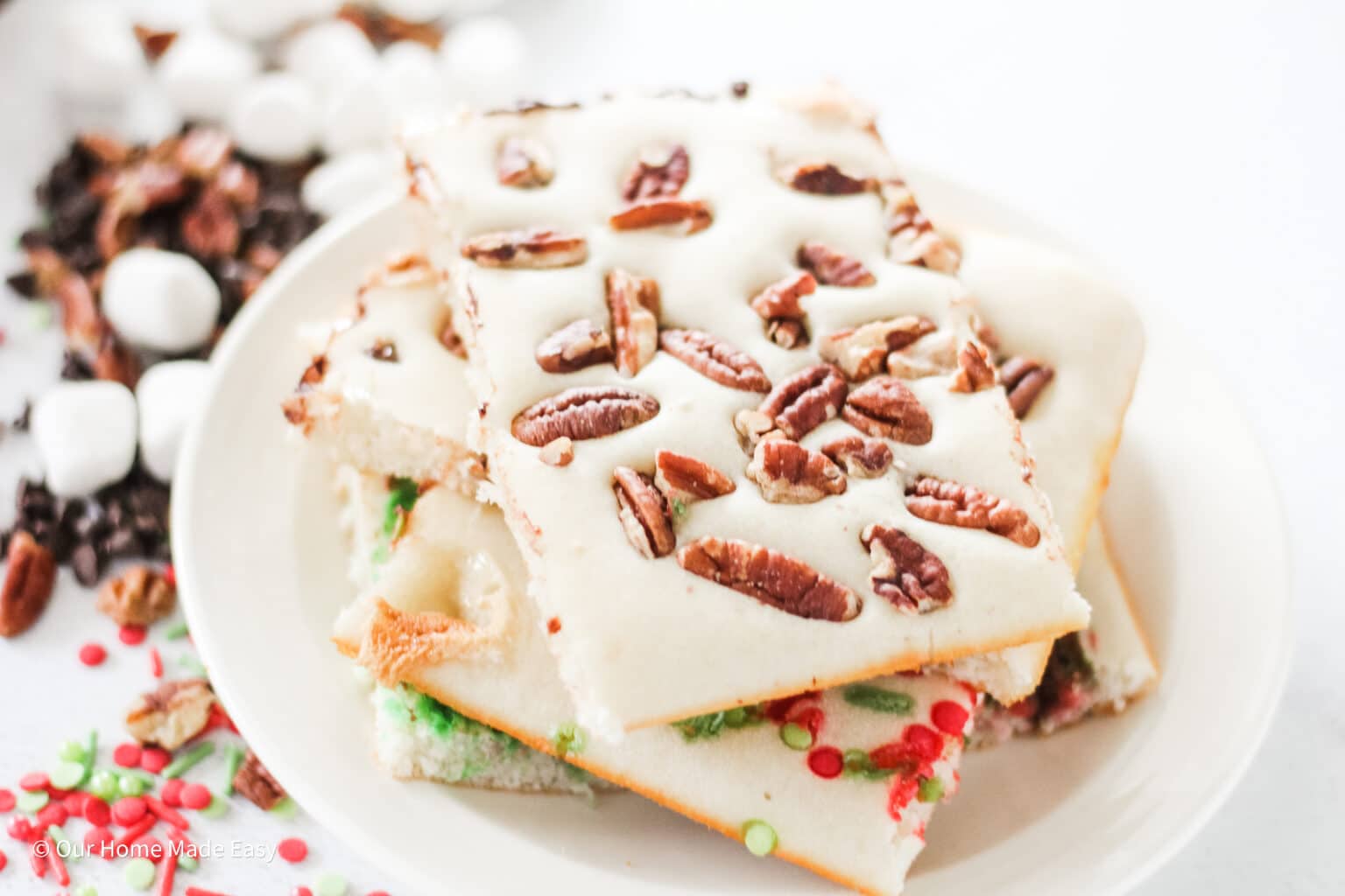 A plate of frosted cookie bars topped with pecans, with marshmallows, chocolate chips, and festive sprinkles scattered nearby.