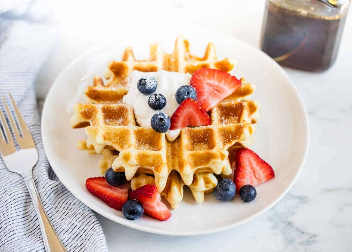 A plate of waffles topped with whipped cream, blueberries, and sliced strawberries, next to a fork and a jar of syrup on a white surface.