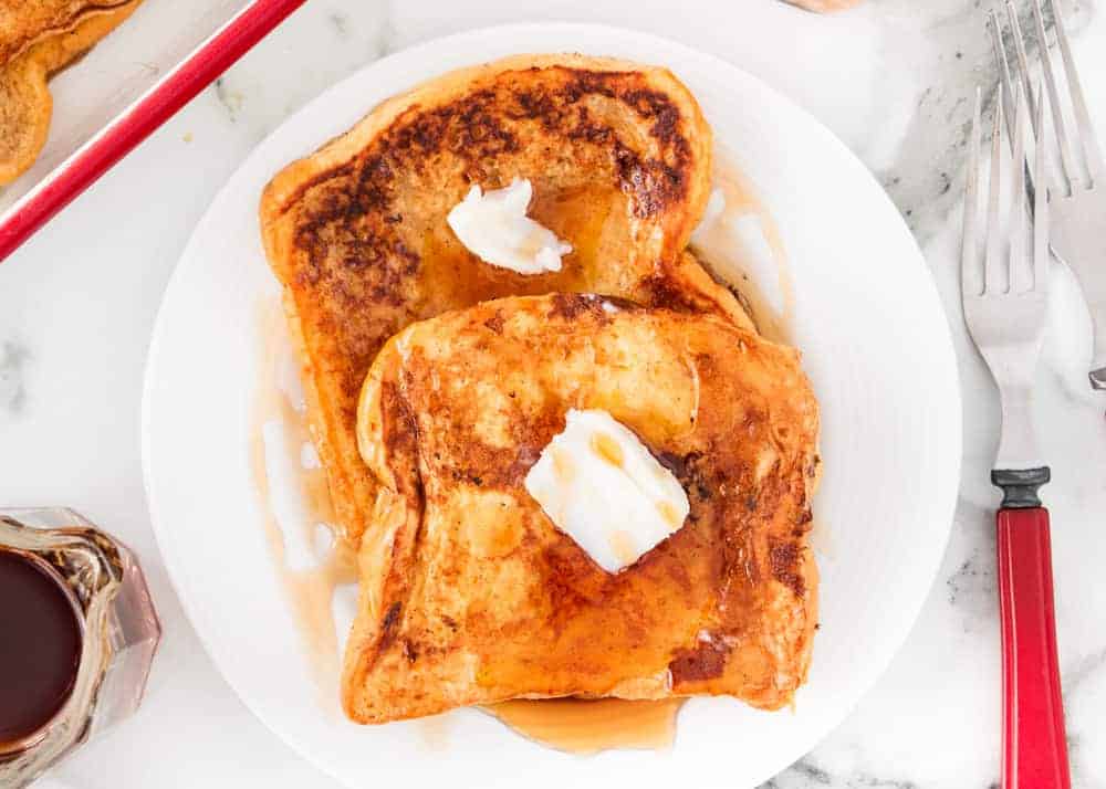 Two slices of French toast topped with butter and syrup on a white plate, with a fork and knife nearby.