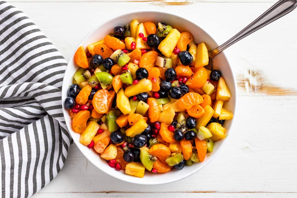 A white bowl filled with a mixed fruit salad including blueberries, kiwi, orange slices, pineapple, and pomegranate seeds sits next to a striped cloth on a white surface.