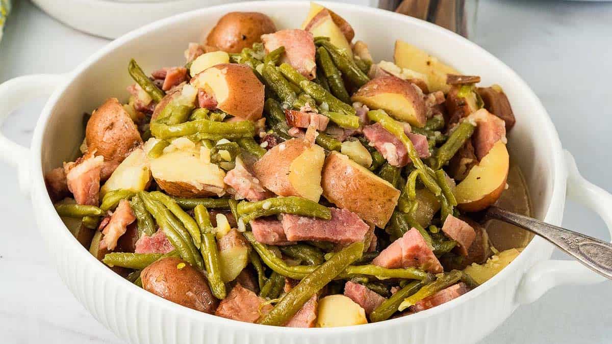 A white bowl filled with cooked green beans, red potatoes, and chunks of ham, with a serving spoon on the side.