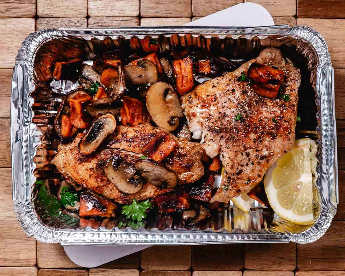 Grilled chicken breast with roasted mushrooms and sweet potatoes, garnished with parsley and lemon wedges, served in a foil container on a wooden surface.