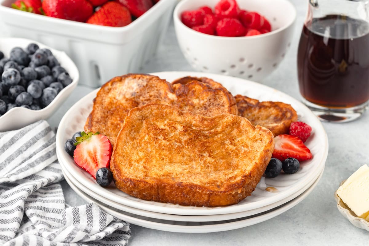Plate of French toast with strawberries, blueberries, and raspberries, next to a jar of syrup, butter, and bowls of fresh berries on a light table.