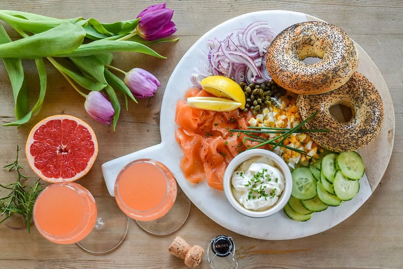 A brunch spread with bagels, smoked salmon, sliced cucumber, red onion, capers, chopped egg, cream cheese, lemon wedges, tulips, a grapefruit half, and two pink drinks on a wooden table.