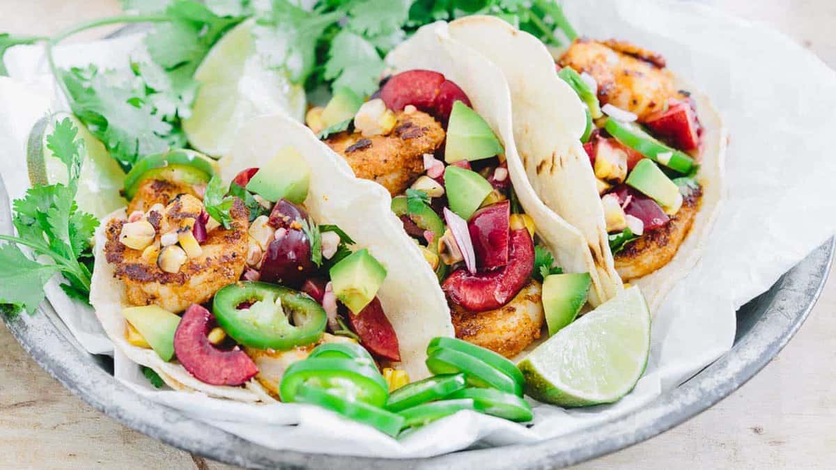 Three soft tacos filled with grilled shrimp, avocado, sliced jalapeรฑos, corn, red onion, and cherries, garnished with cilantro and lime wedges on a plate.