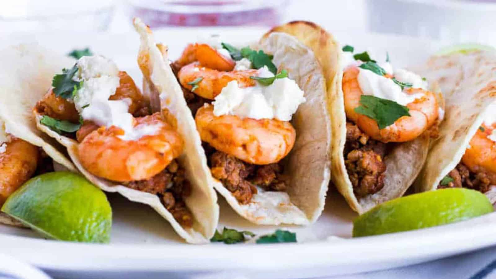 Four shrimp tacos topped with white sauce and cilantro, served on tortillas with lime wedges on a white plate.