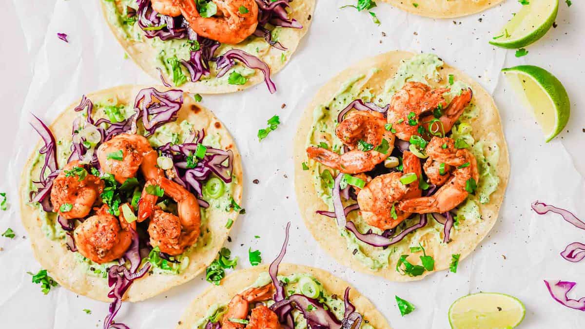 Four shrimp tacos with avocado spread and shredded red cabbage on tortillas, garnished with chopped herbs, are arranged on parchment paper alongside lime wedges.