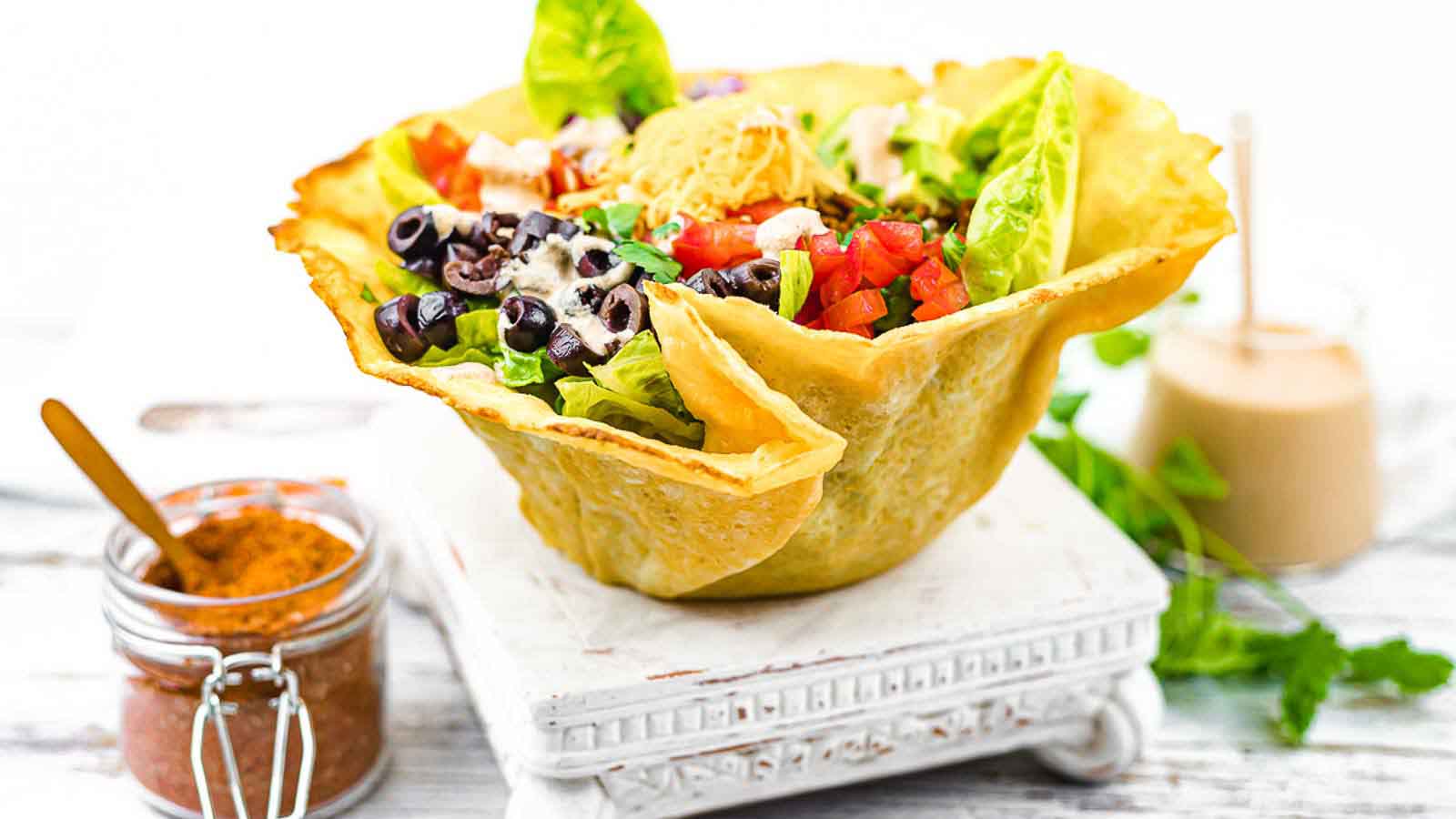 A taco salad served in a crispy tortilla bowl, filled with lettuce, diced tomatoes, black olives, shredded cheese, and dressing. A jar of seasoning and dressing are nearby.