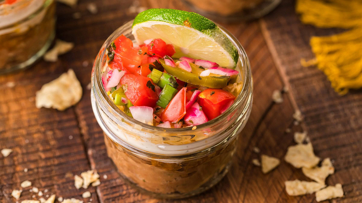 A small glass jar filled with layered dip, topped with diced tomatoes, onions, jalapeรฑos, and a lime slice, sits on a wooden surface with scattered tortilla chips.