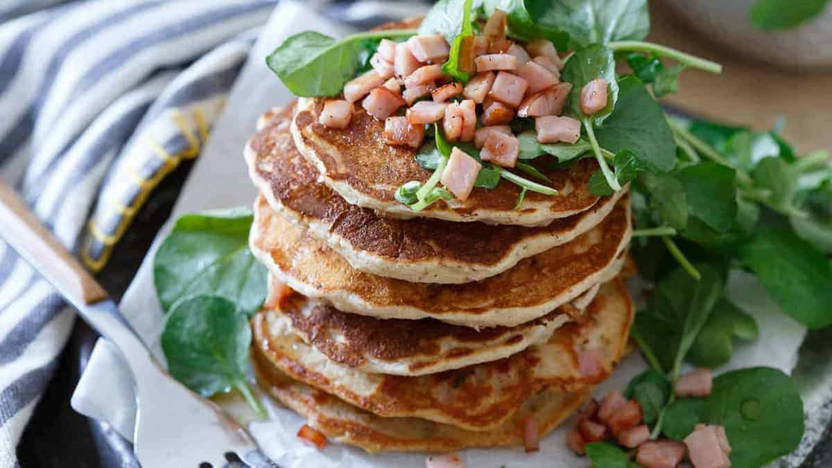 A stack of pancakes topped with diced ham and fresh leafy greens, served on parchment paper with extra greens on the side.