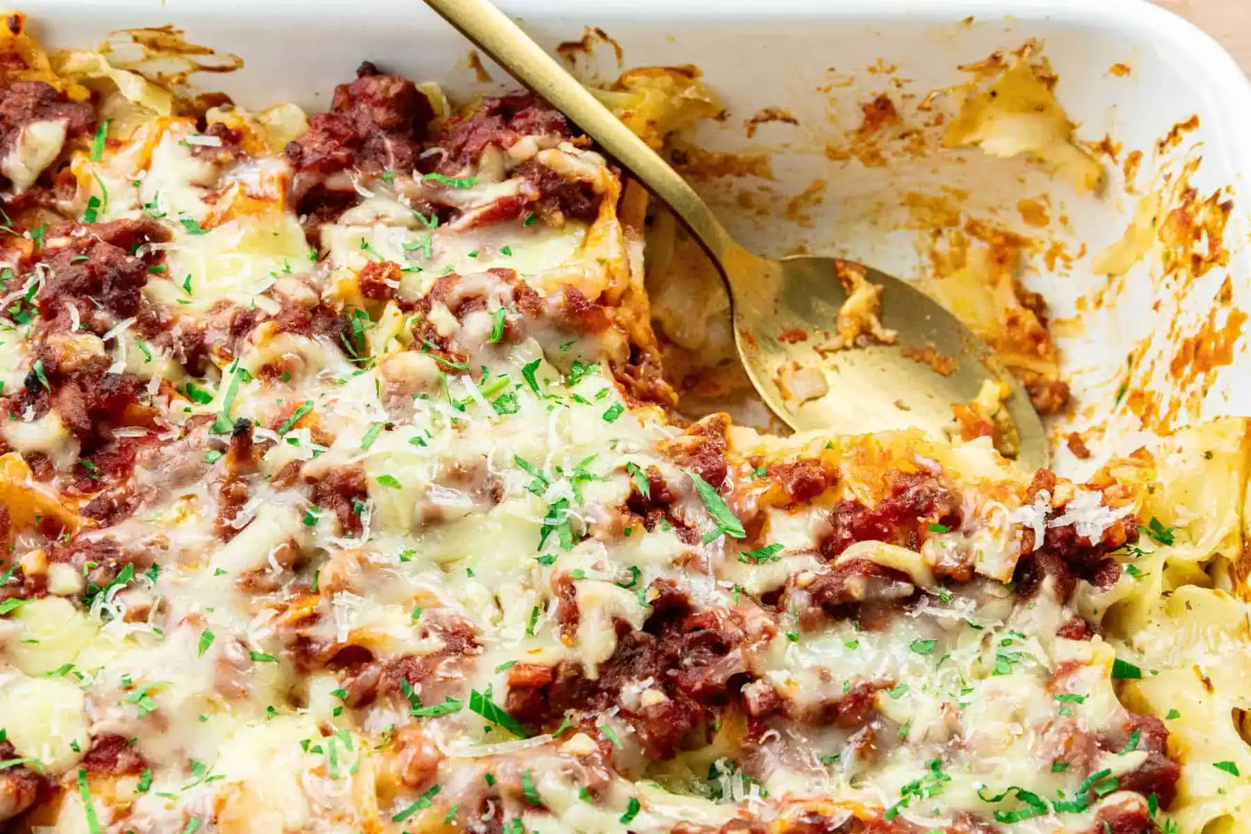 A partially served baked pasta dish topped with melted cheese, tomato sauce, and chopped herbs in a white casserole dish with a serving spoon.