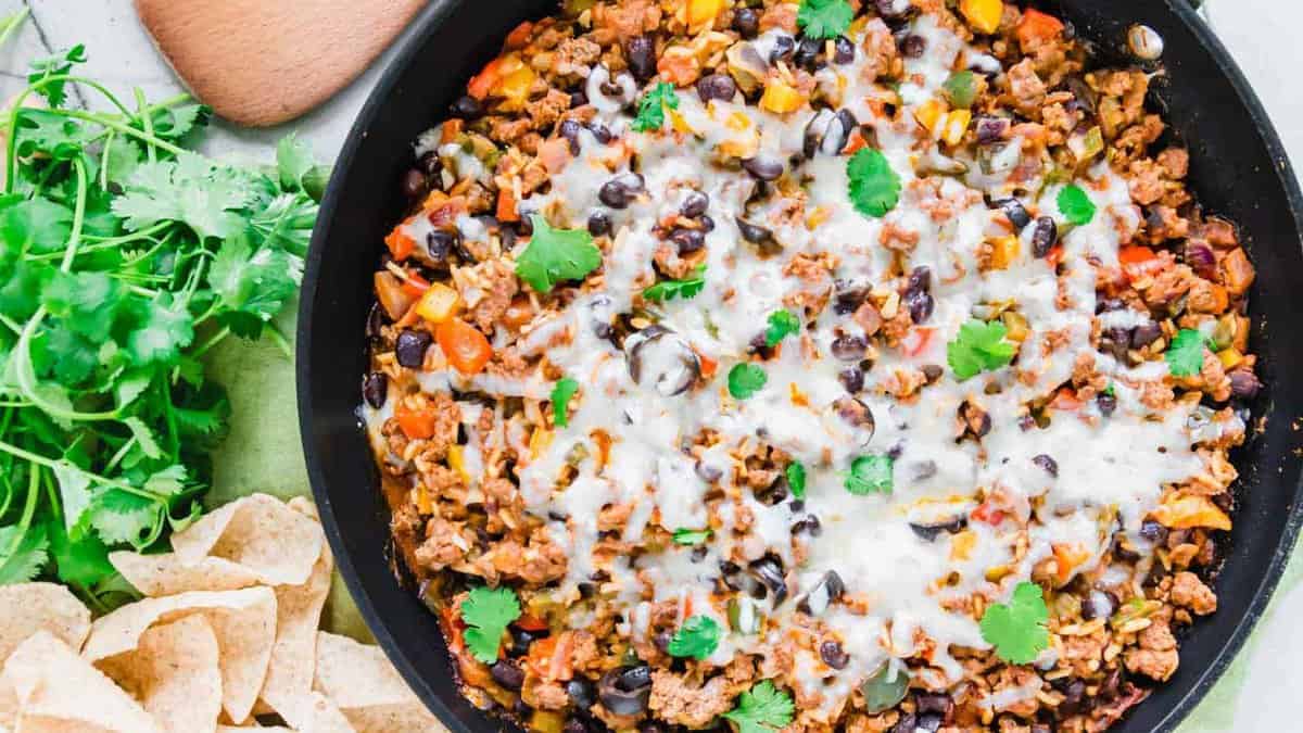 A skillet filled with cooked ground meat, black beans, corn, diced peppers, melted cheese, and cilantro, with tortilla chips and fresh cilantro on the side.