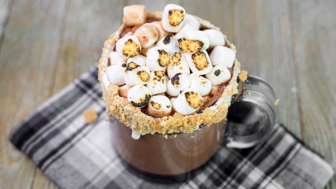 A glass mug of hot chocolate topped with toasted marshmallows and a crushed graham cracker rim sits on a plaid cloth.