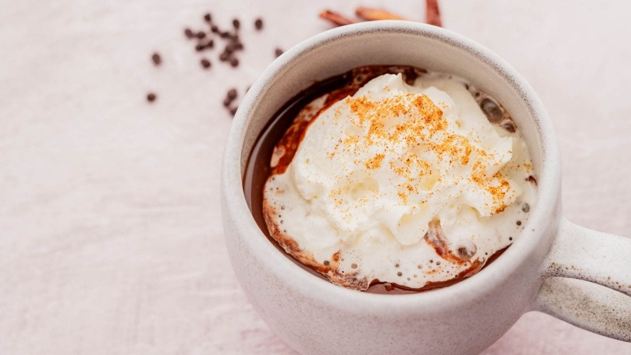 A white mug filled with hot chocolate topped with whipped cream and a sprinkle of cinnamon, placed on a light surface. Chocolate chips are scattered in the background.
