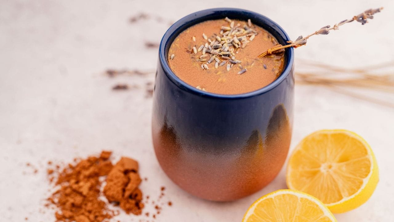 A ceramic mug filled with a brown beverage topped with dried lavender, next to cocoa powder and two lemon halves on a light surface.