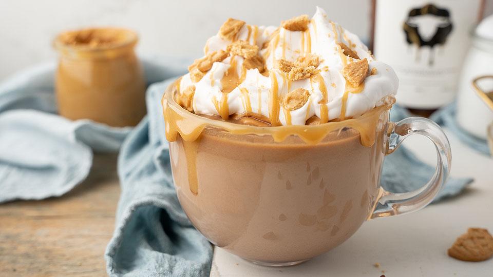 A glass mug filled with a frothy coffee drink topped with whipped cream, caramel drizzle, and crumbled cookies sits on a wooden surface.