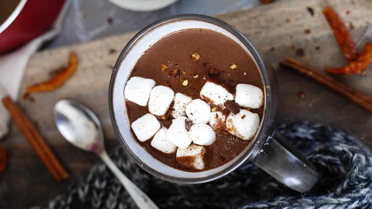 A mug of hot chocolate topped with mini marshmallows sits on a wooden surface, surrounded by cinnamon sticks, a spoon, and a knit scarf.