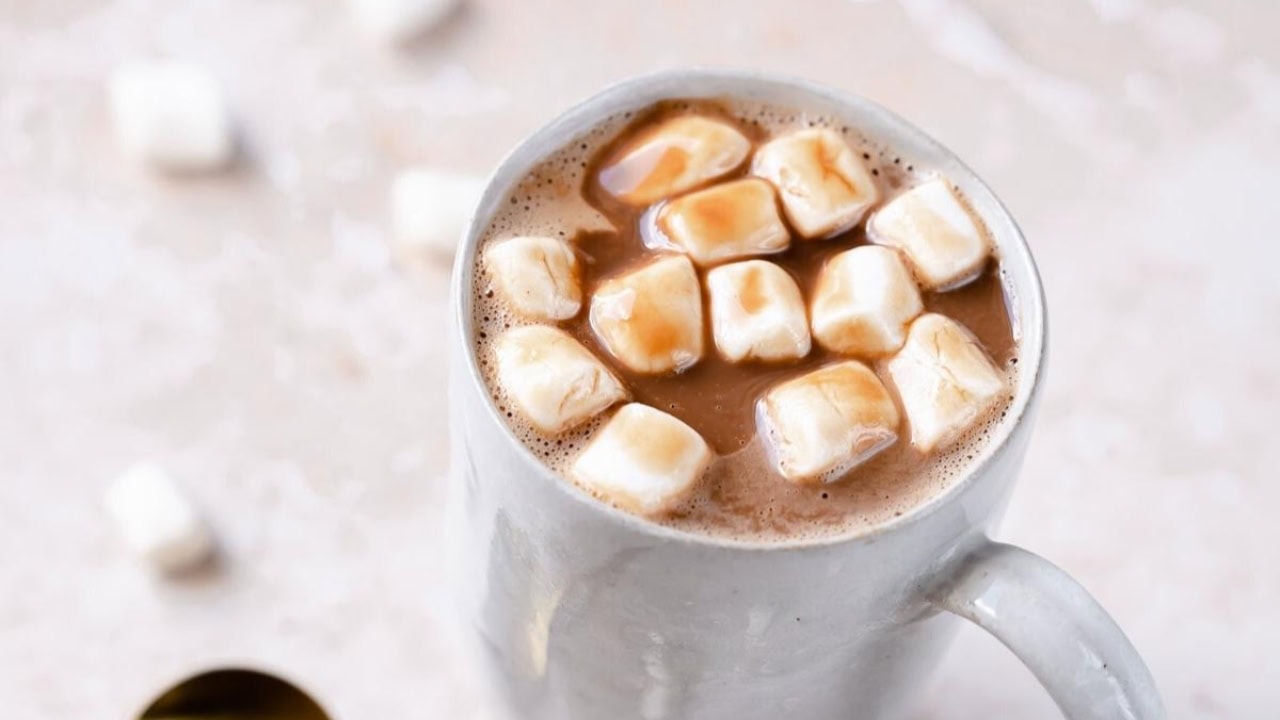 A white mug filled with hot chocolate topped with marshmallows, placed on a light-colored surface with a few marshmallows scattered around.