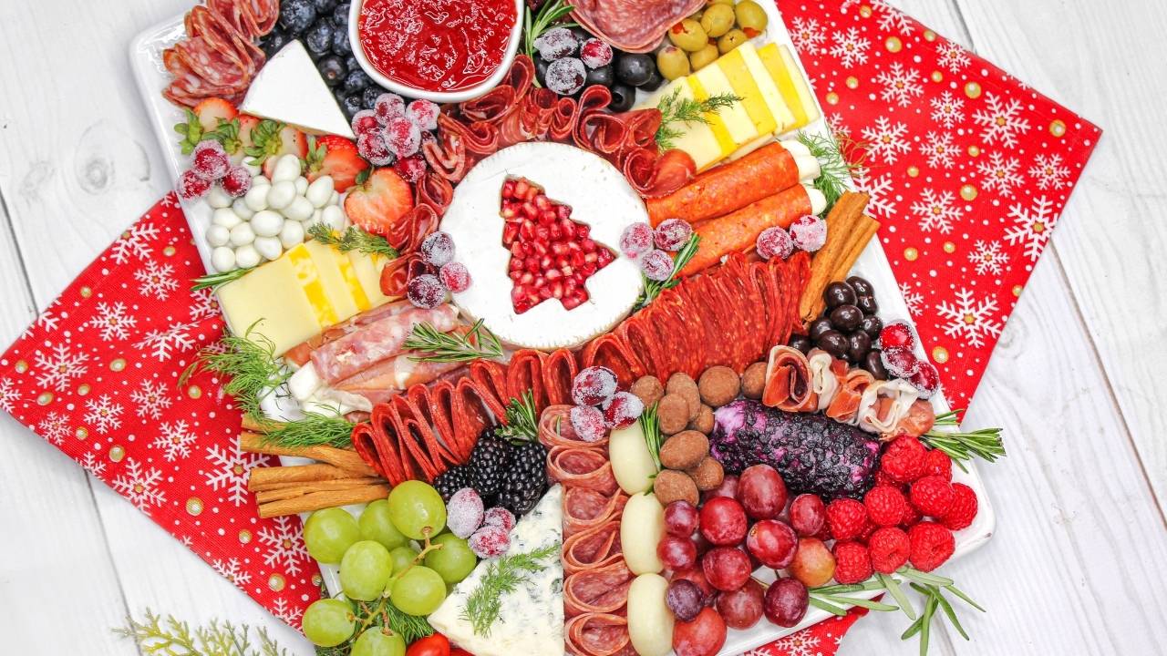 A festive charcuterie board with assorted meats, cheeses, fruits, crackers, and a cheese dip topped with pomegranate seeds shaped like a Christmas tree, on a red snowflake napkin.
