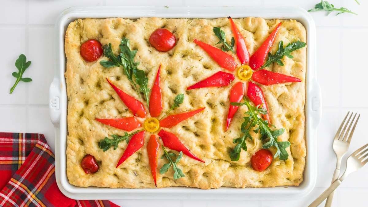 Rectangular focaccia bread topped with red bell pepper slices, cherry tomatoes, yellow tomatoes, and arugula arranged in floral patterns, in a white baking dish on a white surface.
