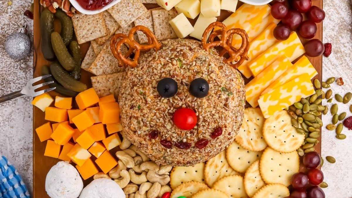 A holiday cheese ball decorated as a reindeer with pretzel antlers, olives and tomato face, surrounded by crackers, cheese cubes, nuts, grapes, pickles, and other snacks.