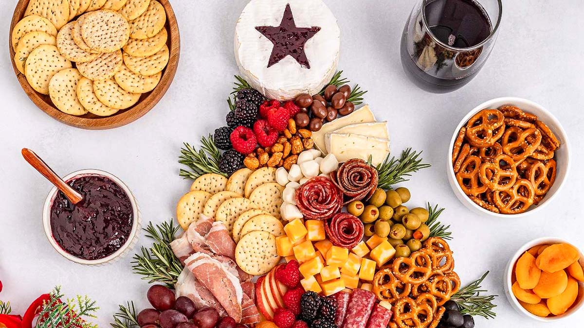 A charcuterie board with assorted cheeses, meats, crackers, pretzels, olives, fruits, jam, dried apricots, and a glass of red wine on a white surface.