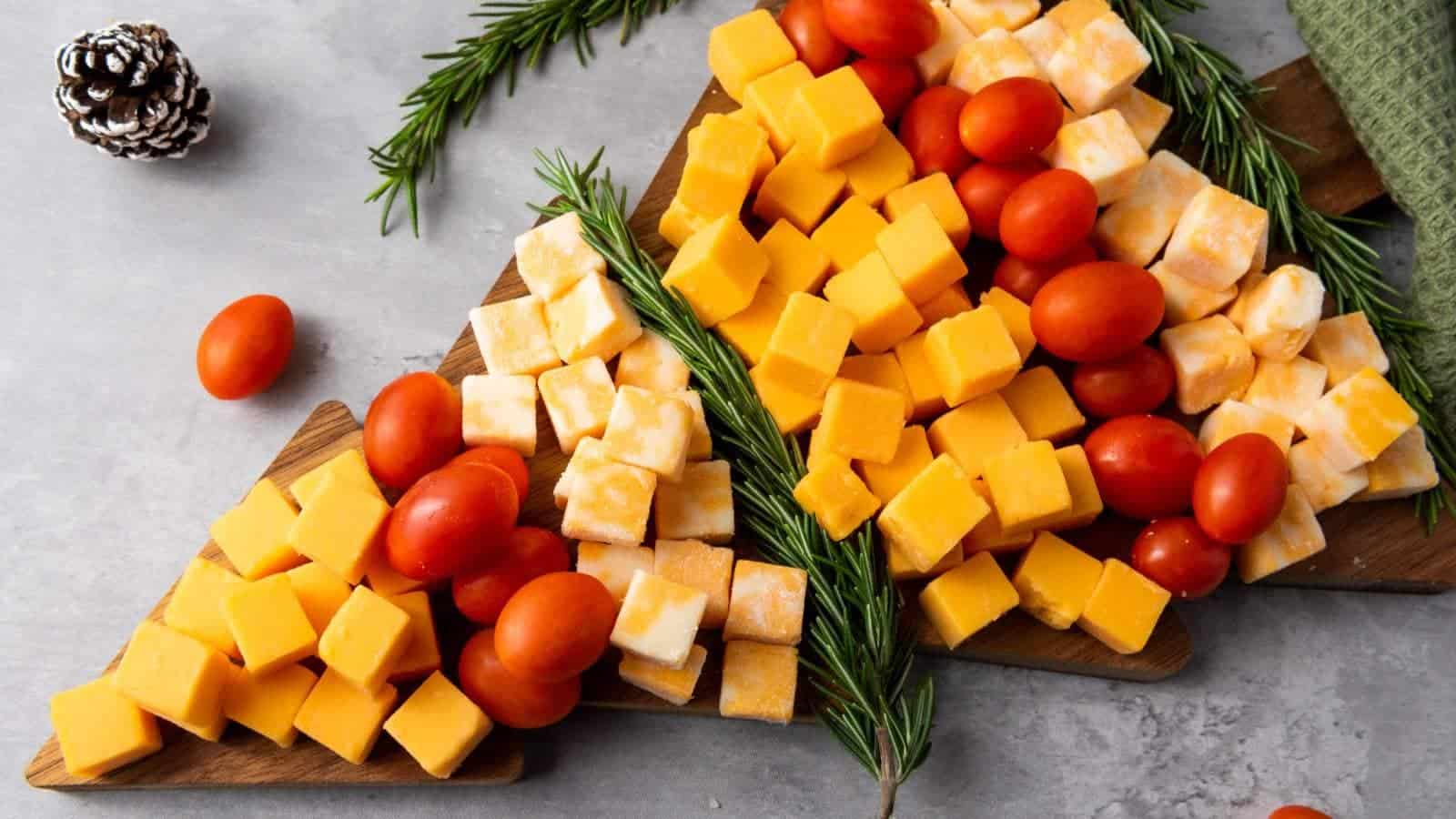 Cubes of cheddar and marbled cheese with grape tomatoes arranged on a wooden board in a triangular shape, garnished with rosemary sprigs.