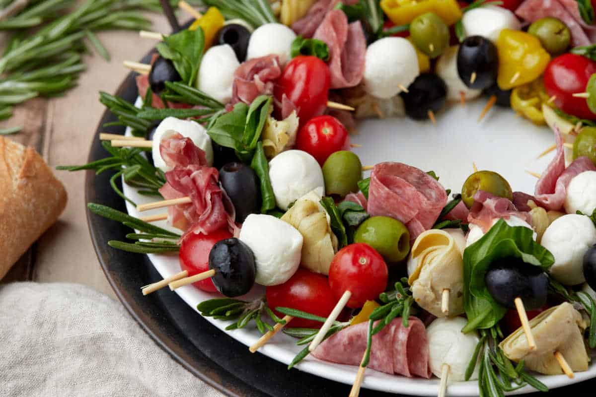 A white plate arranged with skewers holding mozzarella balls, tomatoes, olives, artichokes, salami, and fresh herbs, forming a wreath shape.