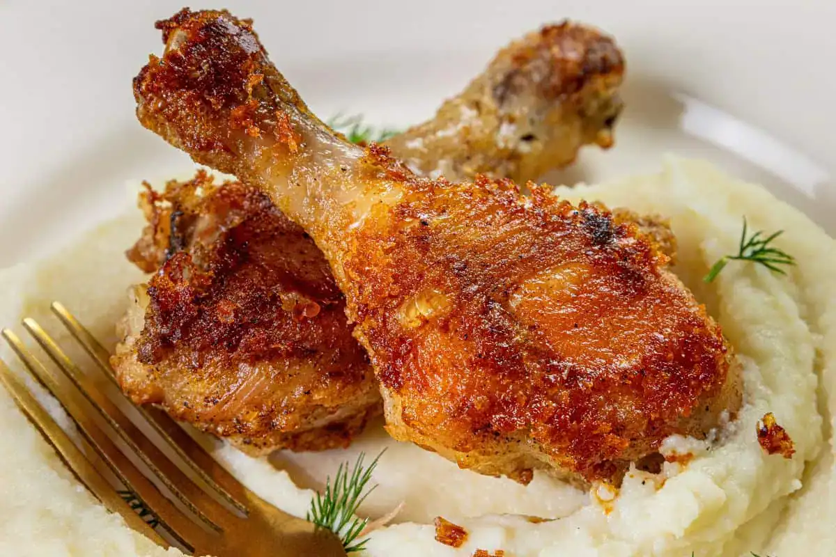 Two crispy fried chicken drumsticks served on a bed of mashed potatoes with a gold fork and a small sprig of dill.