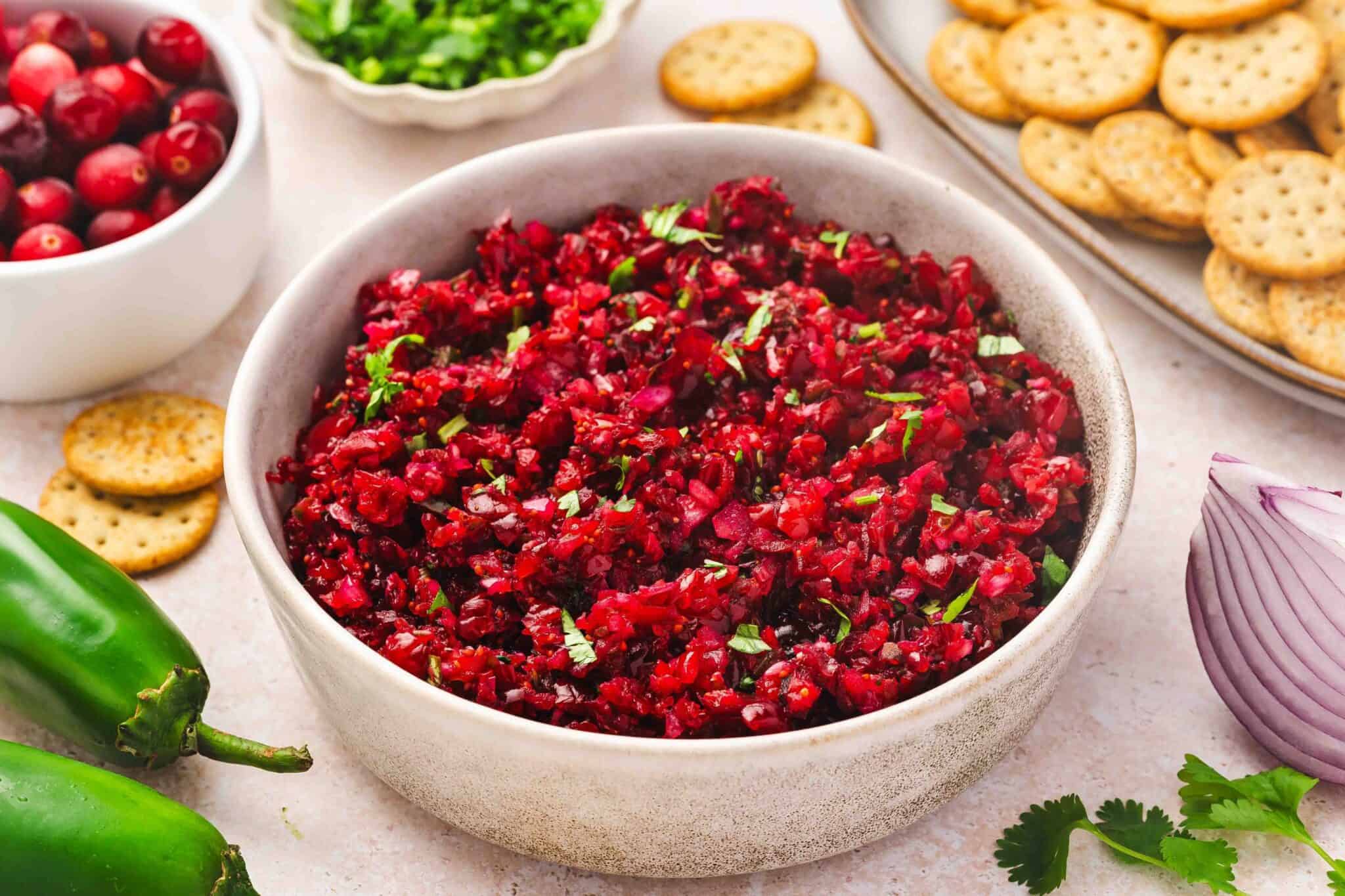 A white bowl filled with cranberry salsa, surrounded by whole jalapeรฑos, crackers, cranberries, chopped herbs, and a slice of red onion.