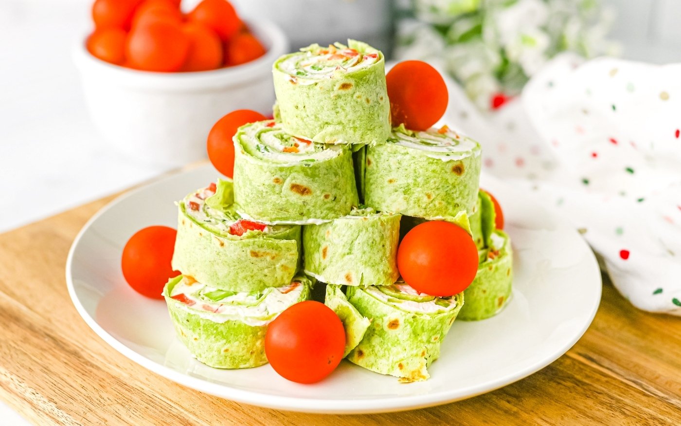 A stack of spinach tortilla pinwheels filled with cream cheese and vegetables, garnished with whole cherry tomatoes on a white plate.