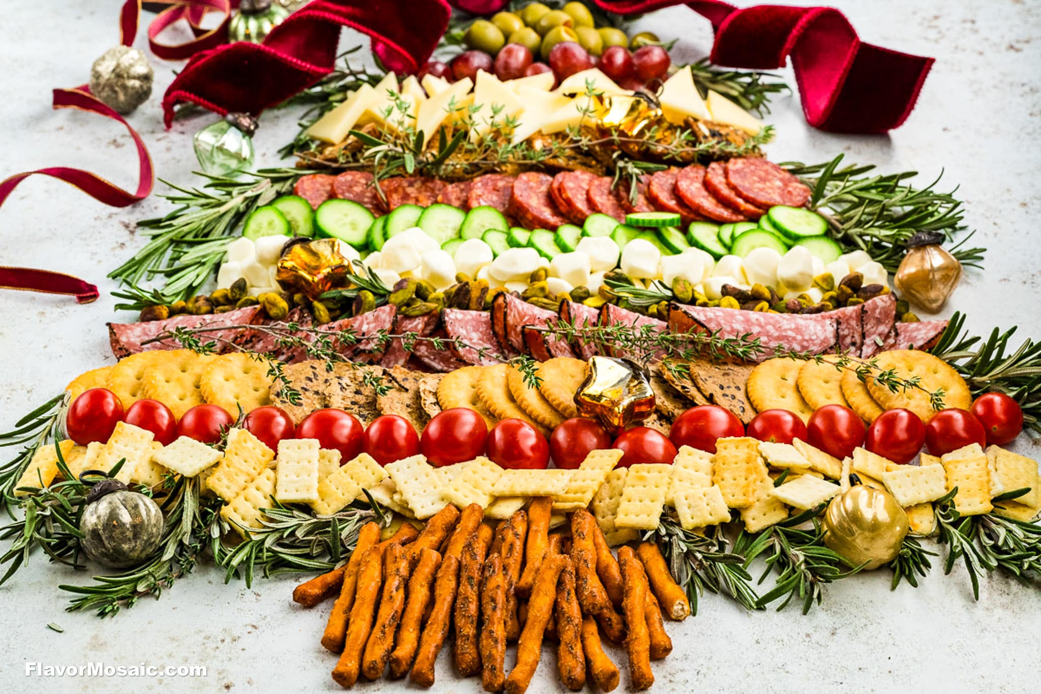A festive charcuterie board arranged in the shape of a Christmas tree, featuring meats, cheeses, crackers, vegetables, and herbs, with holiday decorations and ribbon.