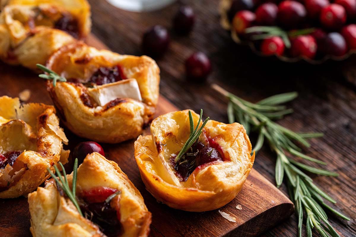 Mini puff pastry tarts filled with cranberry sauce and cheese, garnished with rosemary, on a wooden board with cranberries and herbs in the background.