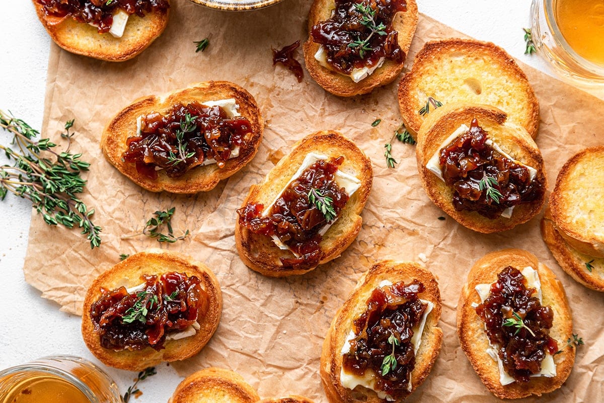 Toasted baguette slices topped with cheese, caramelized onions, and fresh herbs on parchment paper.