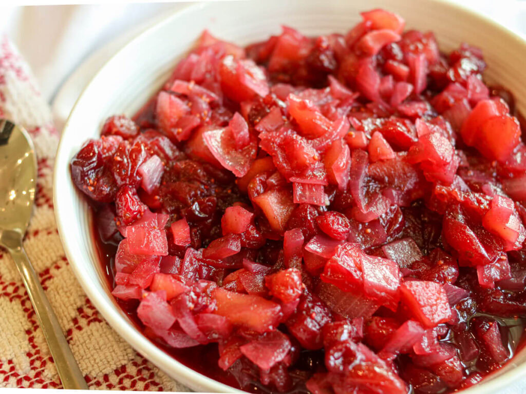 A bowl filled with chunky cranberry relish containing pieces of fruit and onion, placed on a red and white striped cloth next to a spoon.